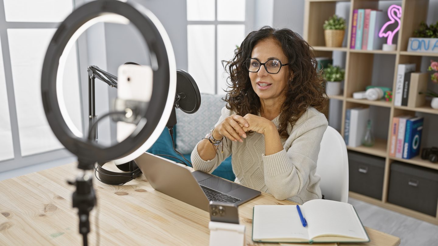 Smiling middle-aged woman recording podcast in a home studio with a laptop, microphone, and ring light